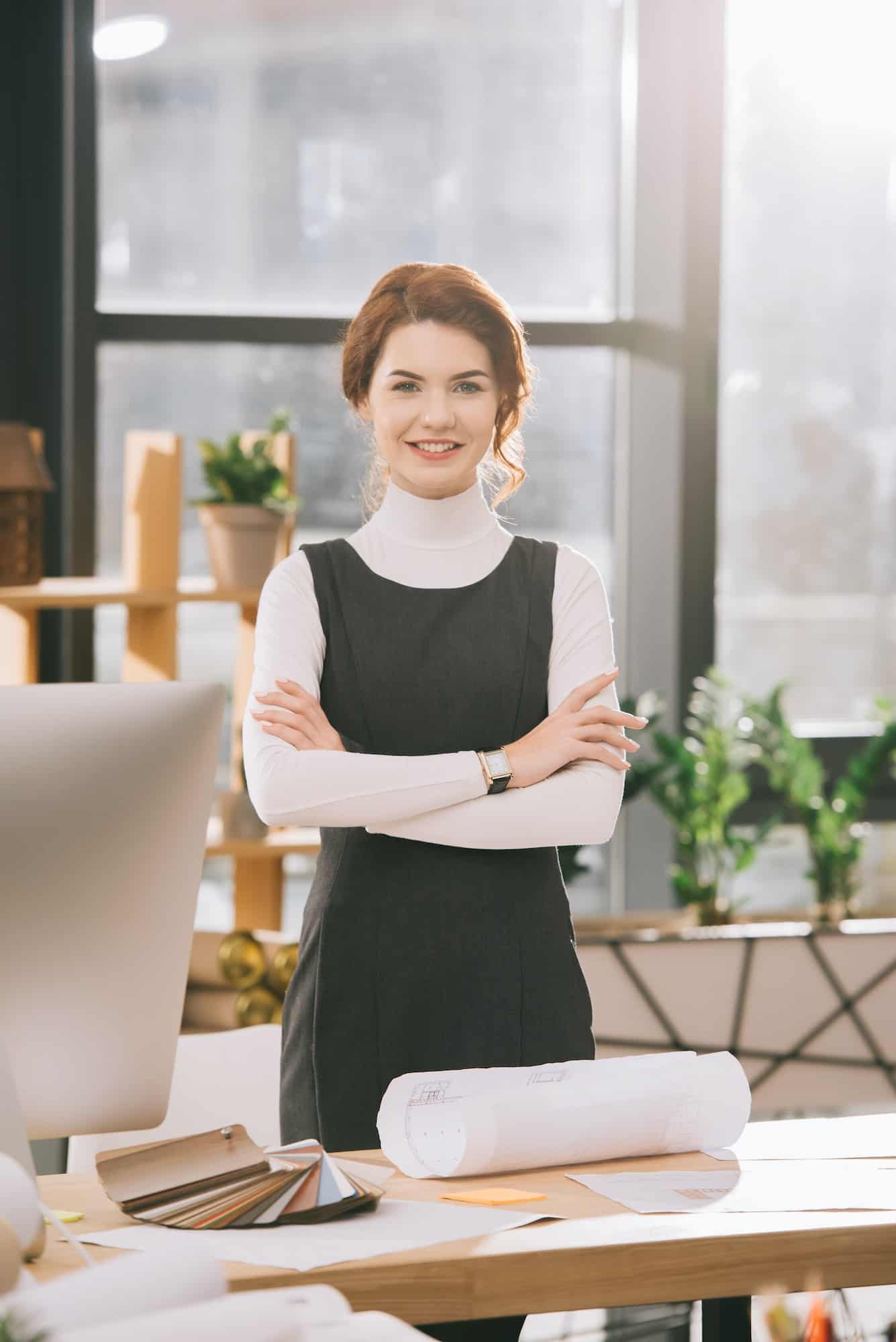 female architect with crossed arms standing at workplace with blueprints